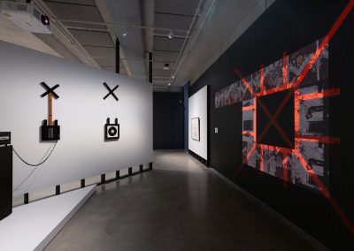 A guitar made by Peter Tyndall is connected to an amplifier and hangs from a wall. On the opposite wall is an artwork with a red cross superimposed on it
