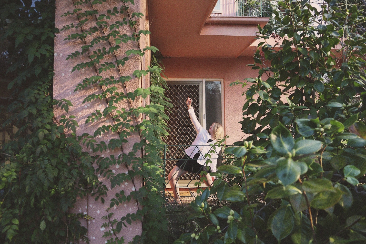 A woman outside on a balcony points to the sky and lunges up with a garden in foreground